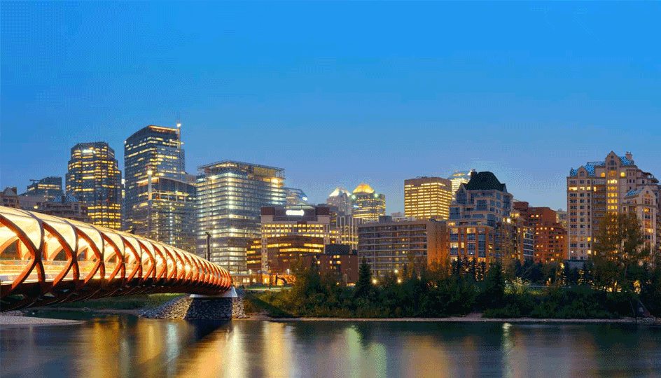 A panoramic view of Calgary, Alberta, Canada at night, featuring the iconic Peace Bridge illuminated against the backdrop of a bustling cityscape